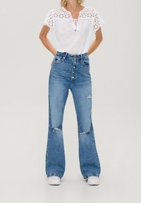 Top bianco a maniche corte con dettagli in pizzo, abbinato a jeans azzurri a vita alta e gamba larga con accenti strappati e chiusura con bottoni.