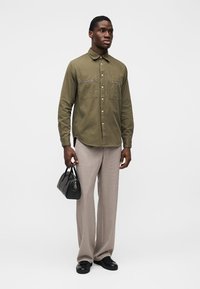 Πουκάμισο σε ελιά πράσινο με κουμπιά και δύο τσέπες στο στήθος με μαύρες λεπτομέρειες, συνδυασμένο με ανοιχτό μπεζ πλισέ παντελόνια και μια μικρή μαύρη τσάντα χειρός.