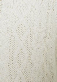 Svetr pletený v krémové barvě s texturovaným, kabelkovým vzorem a složitými detaily, který má měkký a objemný vzhled příze.