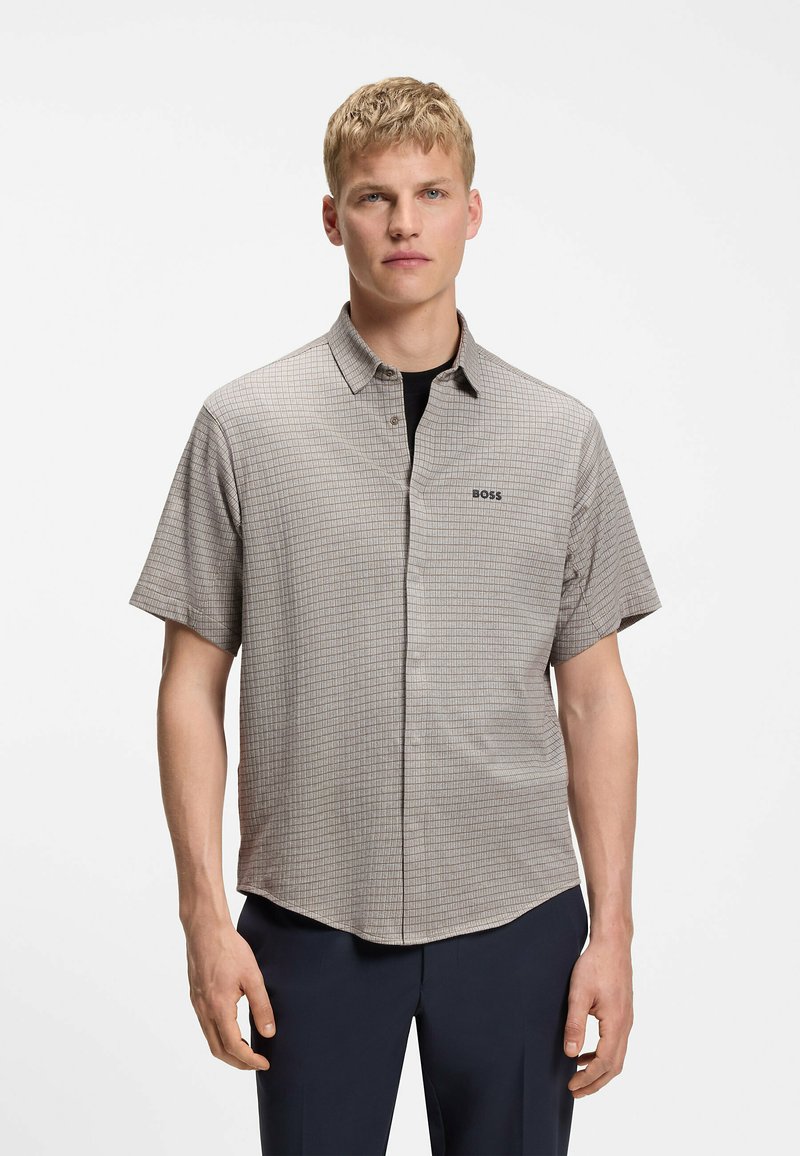 Jeune homme portant une chemise à manches courtes, à carreaux gris clair avec le logo « BOSS » sur la poitrine et un pantalon foncé, debout devant un fond blanc.