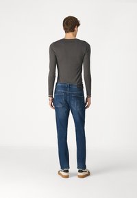 Camisa de manga larga gris y jeans azules vistos desde atrás, mostrando la textura del denim, los bolsillos y un ajuste relajado con zapatillas.