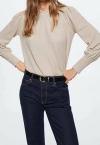 Beige tekstureret langærmet bluse med høj krave og samlede skuldre, parret med mørkeblå jeans og et sort bælte med guldlåse.