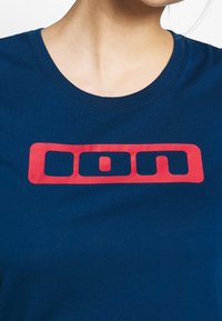 Primer plano de una persona que lleva una camisa azul marino con un logo rectangular rojo que contiene las letras "oom" en el pecho.