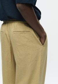 Pantaloni din in bej cu talie elastică, buzunare laterale și un logo brodat subtil pe spate. Textură netedă și croială lejeră.