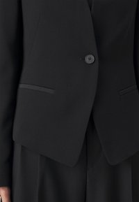 Blazer nero su misura con un singolo bottone, rever a punta e due tasche frontali. Il tessuto ha una texture liscia e un design strutturato.