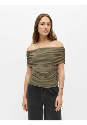 Femme portant un top froncé vert olive à épaules dénudées associé à un jean noir taille haute sur fond blanc uni.