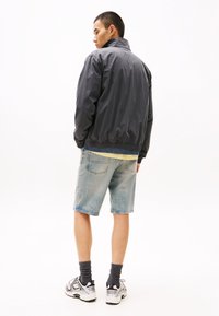 Giacca leggera grigio scuro con colletto rialzato, polsini elastici, indossata sopra una maglietta gialla. Abbinata a pantaloni corti in denim chiaro e scarpe sportive.