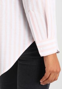Bras et main portant une chemise à manches longues rayée rose et blanche avec un poignet boutonné, reposant à côté d'un pantalon noir.