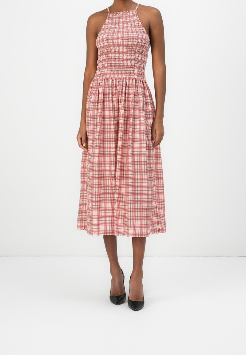 Femme portant une robe midi sans manches à carreaux rouge et blanc avec un corsage smocké et des talons pointus noirs, debout devant un fond blanc.