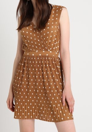 Hnědé šaty bez rukávů s bílými puntíky, plisovanou sukní a zavinovacím výstřihem do V, které nosí stojící žena.