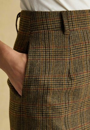 Ruka v kapse hnědých károvaných vlněných kalhot nošených s bílou košilí zastrčenou dovnitř, zobrazující poutka na opasek a detailní texturu látky.