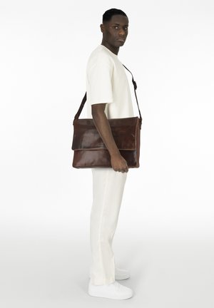 Borsa a tracolla in pelle marrone scuro con design piatto, pattina anteriore e tracolla regolabile, esposta accanto a un modello in tonalità neutre.