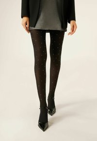 Sorte strømpebukser med en gennemsigtig finish, prydet med små sølvprikker, parret med spidse sorte hæle. Modellen bærer en metallisk grå nederdel.