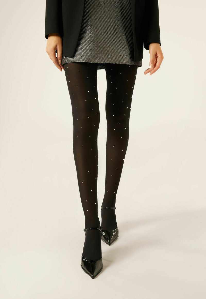 Sorte strømpebukser med en gennemsigtig finish, prydet med små sølvprikker, parret med spidse sorte hæle. Modellen bærer en metallisk grå nederdel.