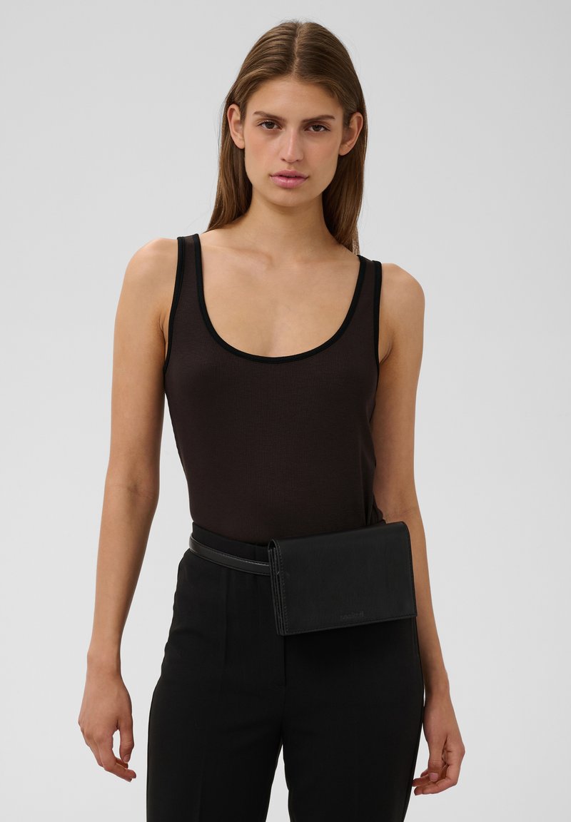 Femme aux cheveux bruns lisses porte un débardeur noir, un pantalon noir et un sac banane noir, debout devant un fond uni clair.