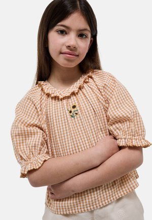 Jeune fille aux longs cheveux bruns porte un chemisier vichy beige et blanc brodé de tournesols, les bras croisés, sur un fond clair uni.