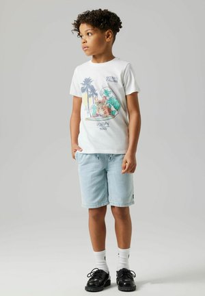 Weißes T-Shirt mit buntem Grafikdesign, das einen Hund und Wellen zeigt, kombiniert mit hellblauen Shorts. Schwarze Schuhe und weiße Socken runden das Outfit ab.