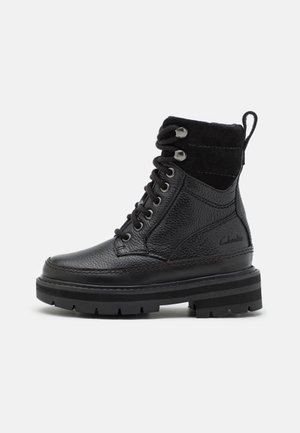 Botte noire en cuir lacée avec col en daim, œillets métalliques, semelle épaisse à rainures et languette à l'arrière, vue de côté.