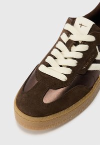 Baskets en daim marron et métal avec lacets crème et semelle en gomme, montrant une étiquette de logo tissée et un détail latéral à rayures.