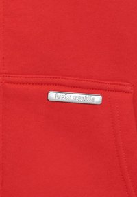 Haut à capuche rouge avec une poche avant, ornée d'une étiquette en métal argenté gravée "bazix republic". Texture lisse et design simple.