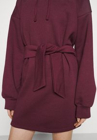 Bordeauxfarbendes, kapuzen Sweatkleid mit einem Bindegürtel. Verfügt über lange Ärmel, eine weiche Textur und eine lockere Passform.