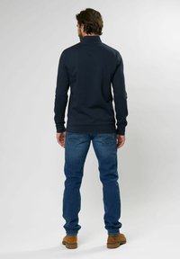 Suéter azul marino con cuello alto, mangas largas, combinado con jeans azules y botas marrones, visto desde atrás sobre un fondo claro.