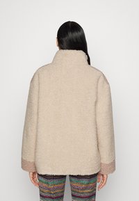 Beige Plüsch-Fleece-Jacke mit hohem Kragen und pinken Akzenten an den Manschetten. Sie hat eine lockere Passform und eine weiche Textur. Getragen über gemusterten Leggings.
