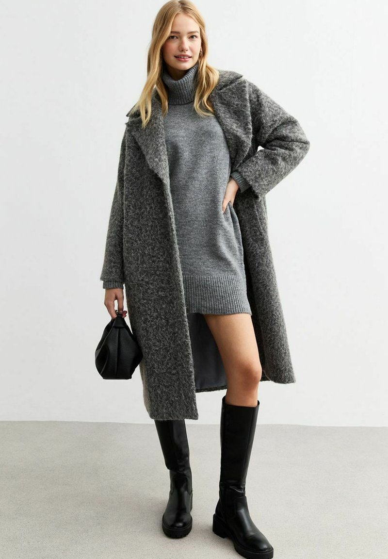 Grauer übergroßer Wollmantel mit strukturiertem Finish, getragen über einem grauen Rollkragenpullover-Kleid. Schwarze knielange Stiefel und eine kleine schwarze Tasche runden den Look ab.