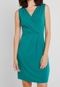 Tealfärgad ärmlös klänning med V-ringning, featuring en draperad byst och slätt, stretchigt tyg. Knälång med lätt uppsamling i midjan.