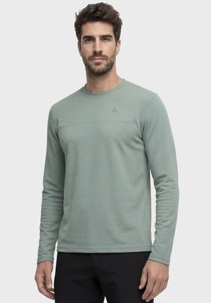 Mann mit kurzen dunklen Haaren und Bart, der ein hellgrünes Langarmhemd und schwarze Hose vor einfarbigem Hintergrund trägt.