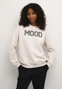 Hellgrauer Pullover mit dem Aufdruck "MOOD" in Schwarz, mit Rundhalsausschnitt, langen Ärmeln und Rippbündchen. Getragen mit schwarzen Hosen.