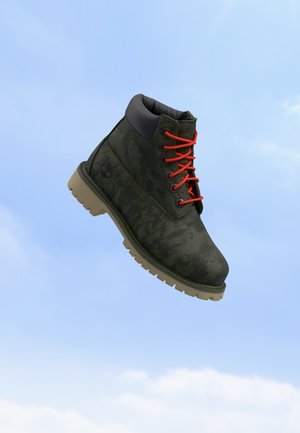 Bota rugosa de color verde oscuro con cordones naranjas y suela beige flotando contra un cielo azul claro con nubes tenues.