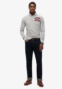 Grijze polo met lange mouwen en hoge kraag, voorzien van rode bedrukte tekst, gecombineerd met donkerblauwe jeans en bruine boot schoenen met opgerolde broekspijpen.