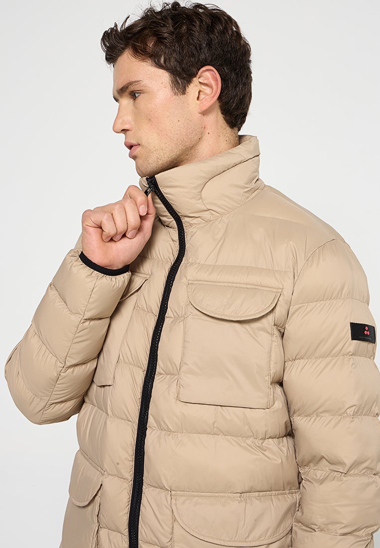 Beige pufferjacka med hög krage, frontdragkedja, två bröstfickor och kontrasterande svart dragkedja. Slät, vadderad textur med en minimalistisk design.