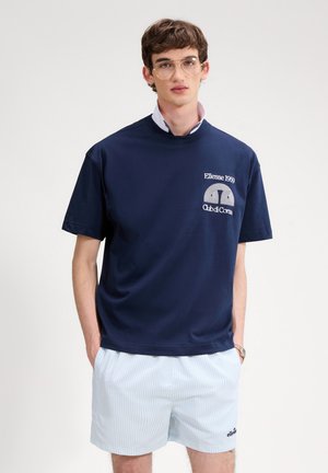 Młody mężczyzna w przezroczystych okularach, granatowej koszulce Ellesse 1959 Club di Corsa oraz jasnoniebieskich, paskowanych szortach z rękami w kieszeniach na białym tle.