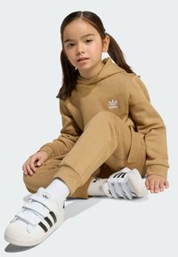 Peščani komplet hupparja in hlač z belim Adidasovim logotipom, dopolnjen z belimi supergami z črnimi črtami in ježkom. Enostaven dizajn, udoben kroj.