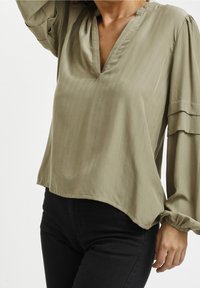 Olivgrüne Bluse mit V-Ausschnitt, langen Puffärmeln und gerafften Manschetten. Weiche Textur mit sanftem Glanz, im lässigen Schnitt.