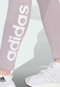 Ljuslila leggings med stort vitt "adidas"-logotyp på sidan, parat med vita mesh-träningsskor med svarta accenter.
