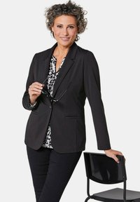 Zwarte blazer met revers, gedragen over een zwart-wit gestippelde top. Model houdt een zonnebril vast en leunt op een zwarte stoel. Slank, aansluitend ontwerp.