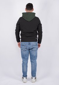 Giacca nera con cappuccio verde, dal design aderente, maniche lunghe e un piccolo logo sulla schiena, abbinata a jeans azzurri chiari.