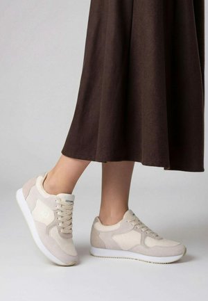 Personne portant des baskets en daim beige avec des semelles blanches et une longue jupe marron, debout sur un fond clair et neutre.