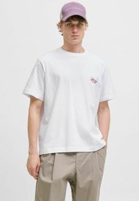 Hvid bomulds t-shirt med korte ærmer, der har et lille rødt broderet logo på brystet. Bärs med beige bukser og en lyse lilla kasket.