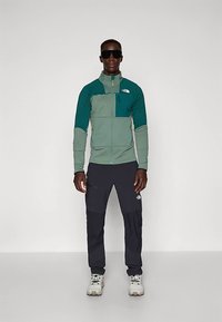 Άνδρας με πράσινη και τιρκουάζ ζακέτα με φερμουάρ, μαύρο παντελόνι σκι, λευκά παπούτσια πεζοπορίας και μεγάλα μαύρα γυαλιά ηλίου, να στέκεται σε λευκό φόντο.