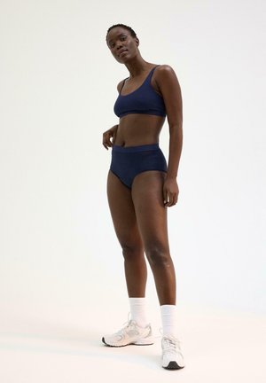 Vrouw staand in een marineblauwe sportbeha en hooggetailleerd ondergoed, met witte sportsokken en sneakers, tegen een effen achtergrond.