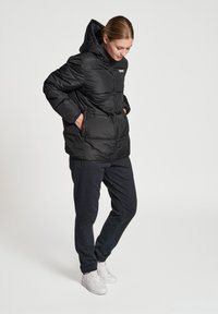 Zwart pufferjack met capuchon, heeft een ritssluiting aan de voorkant, elastische manchetten en zijzakken; gedragen met donkere broek en witte sneakers.