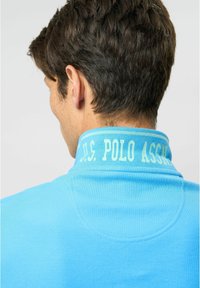 Šviesiai mėlynas polo marškinėlis su tekstūruotu audiniu, turintis apykaklę ir baltu atspaudu "U.S. POLO ASSN." ant nugaros.