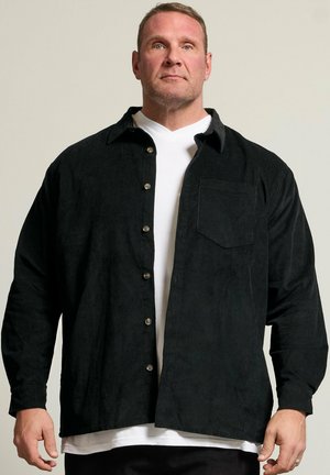 Voksen mand med kort hår iført en åben sort kordfløjls-skjorte over en hvid T-shirt, står mod en ensfarvet baggrund.