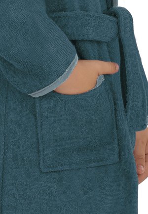 Hand eines Kindes, die in der Tasche eines blauen, strukturierten Bademantels ruht, mit passendem Gürtel, der um die Taille gebunden ist.