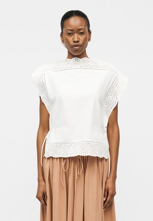 Γυναίκα που φοράει λευκό αμάνικο τοπ από δαντέλα eyelet με κυματιστά τελειώματα και μπεζ πλεκτή φούστα, στέκεται μπροστά σε απλό φόντο.