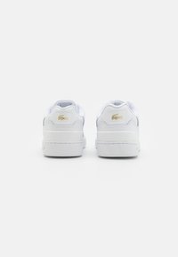 Białe skórzane sneakersy z zaokrąglonym obcasem i wyściełanym kołnierzem, z złotym logo Lacoste z tyłu oraz gumową podeszwą.